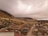 A rural community nestled in a mountainous area under a cloudy sky, featuring dirt roads and various small houses scattered across the landscape. The terrain is hilly and sparsely vegetated with patches of greenery.