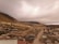 A rural community nestled in a mountainous area under a cloudy sky, featuring dirt roads and various small houses scattered across the landscape. The terrain is hilly and sparsely vegetated with patches of greenery.