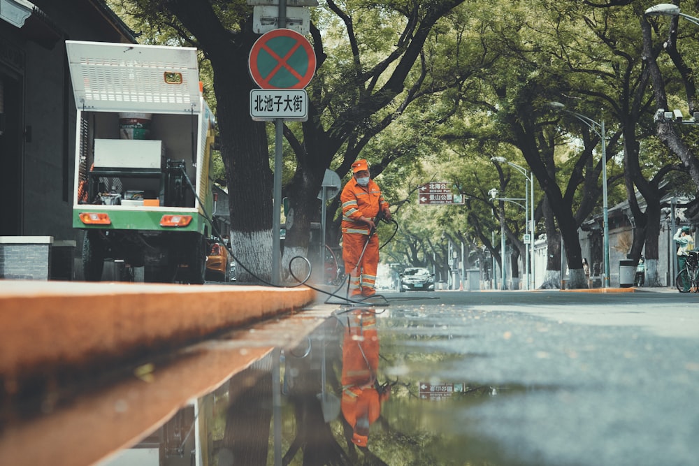 man in orange jacket and black pants standing on sidewalk during daytime