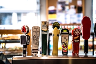 A row of colorful beer taps from various breweries is positioned on a countertop. Each tap handle features a different design and branding, indicating a variety of beers available on draft.