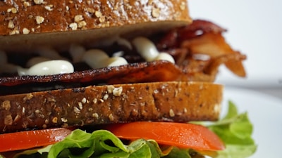 A close-up view of a sandwich made with whole grain bread. Inside, there are crispy bacon strips, slices of fresh tomato, and green lettuce. A white sauce is also visible on the bacon. The sandwich is placed on a white plate, highlighting its fresh ingredients.