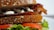 A close-up view of a sandwich made with whole grain bread. Inside, there are crispy bacon strips, slices of fresh tomato, and green lettuce. A white sauce is also visible on the bacon. The sandwich is placed on a white plate, highlighting its fresh ingredients.