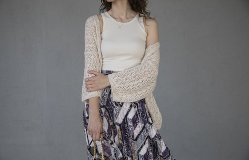 A woman wearing a cream-colored tank top and a crochet cardigan, paired with a patterned skirt. Her arms are loosely crossed, and she stands against a plain gray background. Her wavy hair falls around her shoulders.
