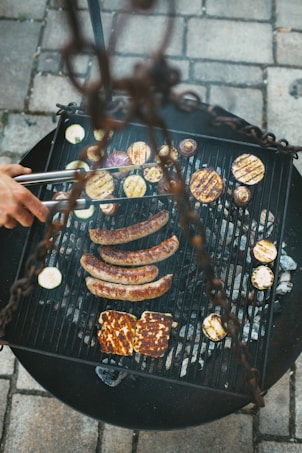 A barbecue grill loaded with sausages and various grilled vegetables, including zucchini slices and possibly pieces of halloumi cheese. A hand holding tongs is seen turning the food, and the scene is set on a stone-tiled patio.