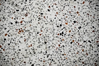 A close-up view of a terrazzo surface featuring a pattern of variously sized and shaped stones embedded in a smooth, light-colored matrix. The stones are predominantly black and brown, creating a speckled effect across the entire surface.