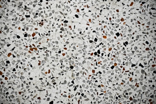 A close-up view of a terrazzo surface featuring a pattern of variously sized and shaped stones embedded in a smooth, light-colored matrix. The stones are predominantly black and brown, creating a speckled effect across the entire surface.