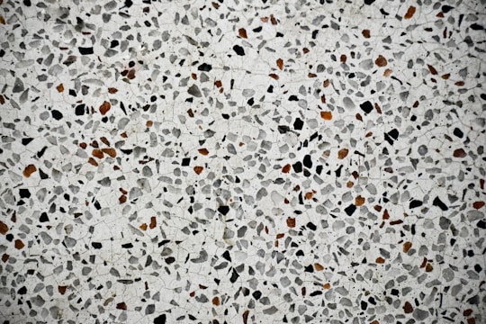A close-up view of a terrazzo surface featuring a pattern of variously sized and shaped stones embedded in a smooth, light-colored matrix. The stones are predominantly black and brown, creating a speckled effect across the entire surface.