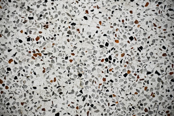 A close-up view of a terrazzo surface featuring a pattern of variously sized and shaped stones embedded in a smooth, light-colored matrix. The stones are predominantly black and brown, creating a speckled effect across the entire surface.