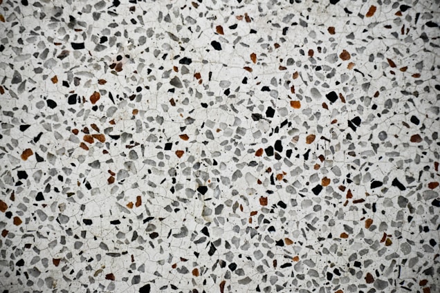 A close-up view of a terrazzo surface featuring a pattern of variously sized and shaped stones embedded in a smooth, light-colored matrix. The stones are predominantly black and brown, creating a speckled effect across the entire surface.