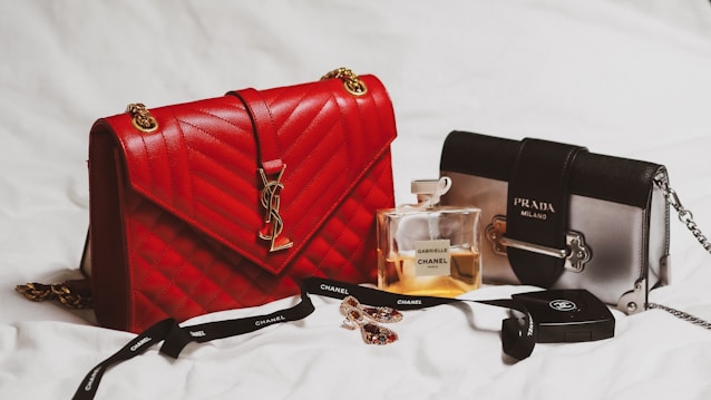 A luxurious arrangement featuring a red quilted handbag with gold chain accents alongside a smaller, two-tone black and gray handbag displaying the Prada logo. A Chanel perfume bottle with a light amber fragrance sits between them, complemented by a black Chanel ribbon and makeup product. A pair of sparkling earrings adds a touch of elegance to this stylish setup.
