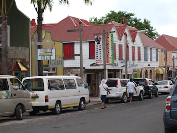 A street scene featuring colorful, historic-looking buildings with red roofs. The storefronts have various signs and are flanked by several parked cars and a couple of pedestrians strolling along the sidewalk. Palm trees are positioned along the street, adding a tropical feel to the setting.