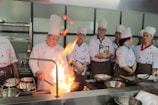 A group of chefs wearing white uniforms and tall chef hats are gathered around a kitchen stove. One chef is actively cooking with a large flame rising from a pan, while others watch attentively. Stainless steel pots and cooking utensils are visible on the stovetop.