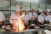 A group of chefs wearing white uniforms and tall chef hats are gathered around a kitchen stove. One chef is actively cooking with a large flame rising from a pan, while others watch attentively. Stainless steel pots and cooking utensils are visible on the stovetop.