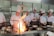 A group of chefs wearing white uniforms and tall chef hats are gathered around a kitchen stove. One chef is actively cooking with a large flame rising from a pan, while others watch attentively. Stainless steel pots and cooking utensils are visible on the stovetop.