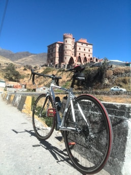 A road bicycle is parked on the side of a road with a striking, castle-like building in the background. The structure features multiple turrets and arched windows, and is situated on a hillside against a clear blue sky. The landscape around is dry with a few shrubs and rolling hills in the distance.
