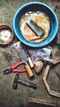 A collection of tools and miscellaneous items are spread out on a textured surface. There is a blue container filled with water and some cleaning cloths at the center. Around it, there are various tools including a pair of red-handled pliers, a hammer, an axe, a chisel, a stone, a partially opened plastic bag with an unidentified object, a small wooden handle tool, several circular metal rings, and a small container filled with nails.