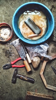 A collection of tools and miscellaneous items are spread out on a textured surface. There is a blue container filled with water and some cleaning cloths at the center. Around it, there are various tools including a pair of red-handled pliers, a hammer, an axe, a chisel, a stone, a partially opened plastic bag with an unidentified object, a small wooden handle tool, several circular metal rings, and a small container filled with nails.
