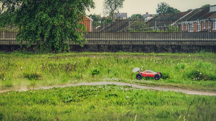 A remote-controlled red car is moving across a lush green field. In the background, there is a line of residential houses with sloped roofs, and a large tree is visible near a fence.