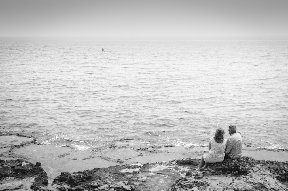 grayscale photo of woman sitting on rock by the sea