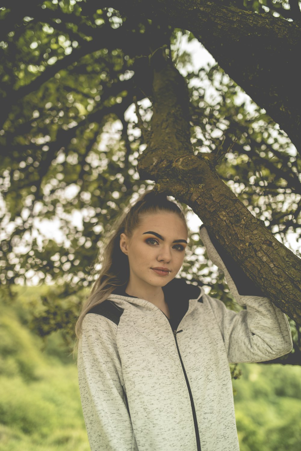 woman in gray hoodie standing beside brown tree during daytime