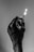 A hand covered in dark paint or ink holds a lit matchstick against a plain background. The matchstick emits a small, bright flame.