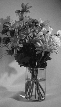 A grayscale photograph featuring a bouquet of various flowers arranged in a transparent glass vase. The flowers have different shapes and petal arrangements, creating a textured and layered composition. The background is plain, casting subtle shadows that add to the depth of the image.