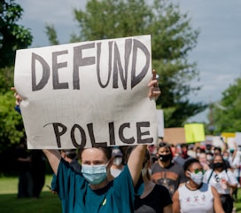 A group of people participating in a protest. The main individual in the foreground is holding a large sign that reads 'DEFUND POLICE'. Many participants are wearing face masks, and the gathering is taking place outdoors, surrounded by greenery.