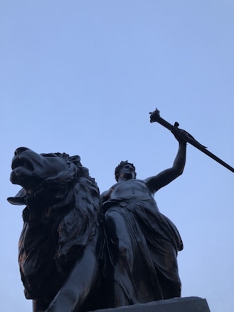 A large bronze statue featuring a powerful lion and a human figure holding a scepter. The perspective is from below, emphasizing the grandeur and dominance of the figures against a clear blue sky.