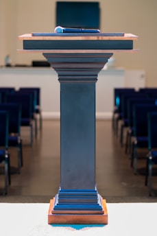 A microphone rests on top of a wooden podium with a smooth, dark finish. The podium stands in a room set up with rows of blue chairs, facing a small stage or presentation area. The background includes office equipment, suggesting a formal setting like a conference room or lecture hall.