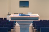 A wooden podium is positioned in a room with rows of empty seating, and a microphone rests on top. In the background, there is a blurred monitor on the wall, enhancing the setting's focus on public speaking or presentations.
