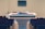 A wooden podium is positioned in a room with rows of empty seating, and a microphone rests on top. In the background, there is a blurred monitor on the wall, enhancing the setting's focus on public speaking or presentations.