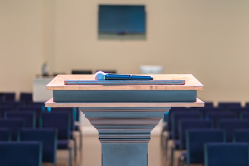 A wooden podium is positioned in a room with rows of empty seating, and a microphone rests on top. In the background, there is a blurred monitor on the wall, enhancing the setting's focus on public speaking or presentations.