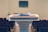 A wooden podium is positioned in a room with rows of empty seating, and a microphone rests on top. In the background, there is a blurred monitor on the wall, enhancing the setting's focus on public speaking or presentations.