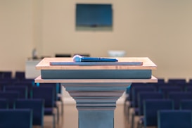 A wooden podium is positioned in a room with rows of empty seating, and a microphone rests on top. In the background, there is a blurred monitor on the wall, enhancing the setting's focus on public speaking or presentations.