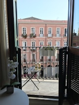 A balcony view from a room showing a small table with a white orchid plant inside and two bottles on a wooden chair outside. The background features an elegant pink building with white-trimmed windows and flower boxes under a clear blue sky.