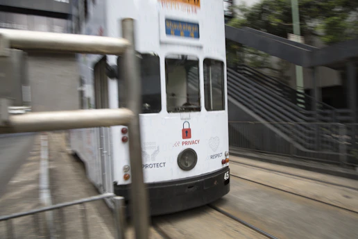 A white tram is moving along its tracks in an urban environment. The tram features various text and graphics, including symbols related to privacy and protection. There are steel barriers in the foreground, and an outdoor staircase with metal railings is visible in the background.