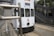 A white tram is moving along its tracks in an urban environment. The tram features various text and graphics, including symbols related to privacy and protection. There are steel barriers in the foreground, and an outdoor staircase with metal railings is visible in the background.