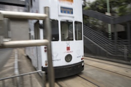 A white tram is moving along its tracks in an urban environment. The tram features various text and graphics, including symbols related to privacy and protection. There are steel barriers in the foreground, and an outdoor staircase with metal railings is visible in the background.