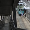 A green double-decker tram is stationary at a tram stop, with advertisements for travel to the Philippines. Two people are crossing the tram tracks in front of the vehicle. The perspective is from a staircase leading down to the street level.