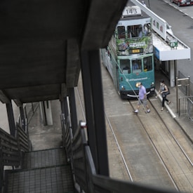 A green double-decker tram is stationary at a tram stop, with advertisements for travel to the Philippines. Two people are crossing the tram tracks in front of the vehicle. The perspective is from a staircase leading down to the street level.