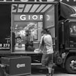 A black and white photograph captures a delivery scene where a man is unloading or organizing parcels from a truck. The back of the truck is open, revealing a variety of packages. In the background, there's a store with a prominent sign displaying 'GIOR' and numerous clothing items and mannequins visible inside. The street is busy, with another cart positioned near the truck.