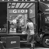 A black and white photograph captures a delivery scene where a man is unloading or organizing parcels from a truck. The back of the truck is open, revealing a variety of packages. In the background, there's a store with a prominent sign displaying 'GIOR' and numerous clothing items and mannequins visible inside. The street is busy, with another cart positioned near the truck.