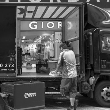 A black and white photograph captures a delivery scene where a man is unloading or organizing parcels from a truck. The back of the truck is open, revealing a variety of packages. In the background, there's a store with a prominent sign displaying 'GIOR' and numerous clothing items and mannequins visible inside. The street is busy, with another cart positioned near the truck.