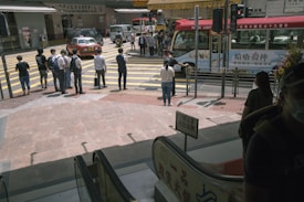 A busy urban street scene with people waiting at a crosswalk. A red minibus and a taxi are visible, along with several pedestrians standing at the edge of the sidewalk. The background includes various storefronts and signage, capturing a typical city atmosphere.
