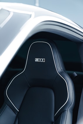 A sleek and modern car seat with black leather upholstery, featuring white stitching and a bold logo on the headrest. The seat is positioned within a partially open car door, revealing a stylish and futuristic design.