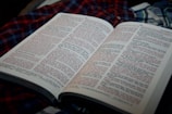 An open book displaying text in two columns, with some portions of the text highlighted in red. The book is placed on a checkered fabric, predominantly in shades of red and blue.