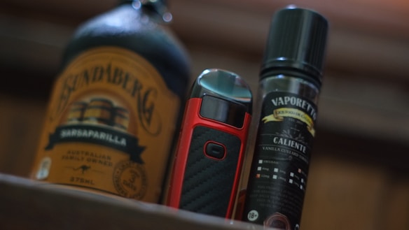 A bottle of Bundaberg Sarsaparilla is positioned alongside a red and black vape device and a Vaporette e-liquid bottle. The Bundaberg bottle features a brown label with vintage-style branding. The vape device has a sleek design with a prominent button, and the e-liquid bottle displays a detailed label with various texts and graphics, suggesting a vanilla custard flavor.