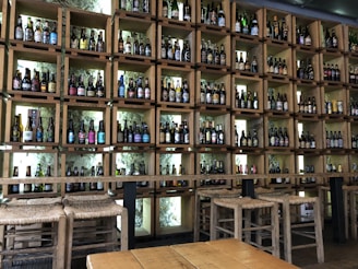 Wooden shelves lined with a wide variety of beer bottles, arranged in a grid pattern. The shelves are backlit, highlighting the labels and shapes of the bottles. A wooden table and rustic stools in the foreground suggest a cozy, artisanal setting.