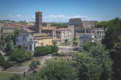 A historic cityscape featuring ancient Roman architecture, including a multi-tiered structure resembling the Colosseum and several other classical buildings. The scene is surrounded by lush greenery and dense clusters of modern and ancient buildings.