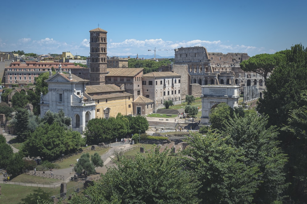 A historic cityscape featuring ancient Roman architecture, including a multi-tiered structure resembling the Colosseum and several other classical buildings. The scene is surrounded by lush greenery and dense clusters of modern and ancient buildings.